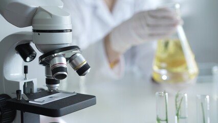 Microscope is near a scientist, wearing a lab coat and white protective gloves, and holding beaker with a yellow oily liquid in laboratory setting, close up. Medicine and science concept