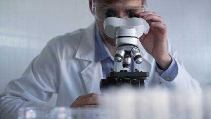 Male scientist wearing lab coat and protective glasses using microscope doing research in modern...