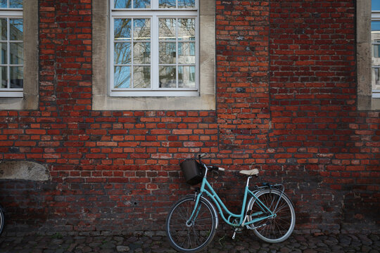A charming turquoise vintage bicycle with a black basket leans against an old red brick wall with white-framed windows.
