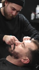 Professional barber carefully shaving client's beard with straight razor, applying gel and using...