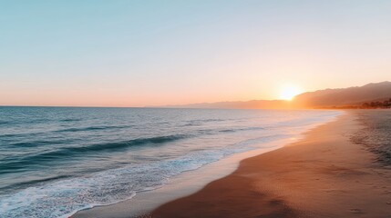Fototapeta premium Waves gently crash on the sandy beach as the orange sun sets over the ocean, illuminating the surrounding mountains in a tranquil setting