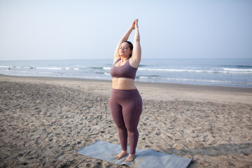 plus size girl does yoga on  the sea beach to lose weight and keep fit. The woman is wearing sports clothes, a purple top and leggins.