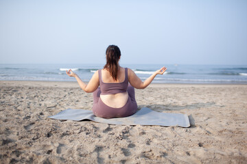 plus size girl does yoga on  the sea beach to lose weight and keep fit. The woman is wearing sports...