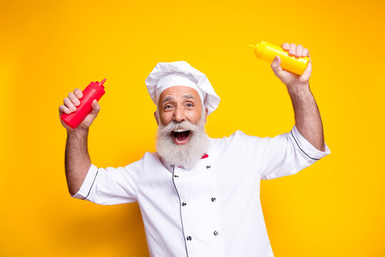 Happy senior chef in uniform holding mustard and ketchup bottles on vibrant yellow backdrop