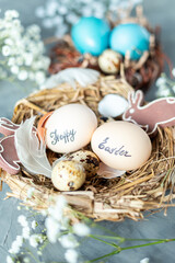 Two chicken eggs with the inscription Happy Easter in a nest of dry grass with feathers, rabbits and gypsophila branches on a light blue background.