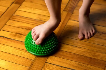 A child's bare feet stand on a green spiky massage ball on a wooden floor, promoting balance and relaxation.