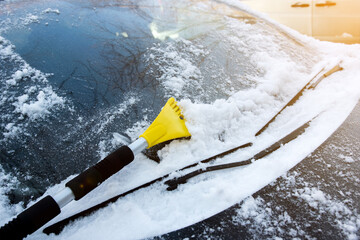 Yellow ice scraper clearing snow from car windshield, sunny winter morning.