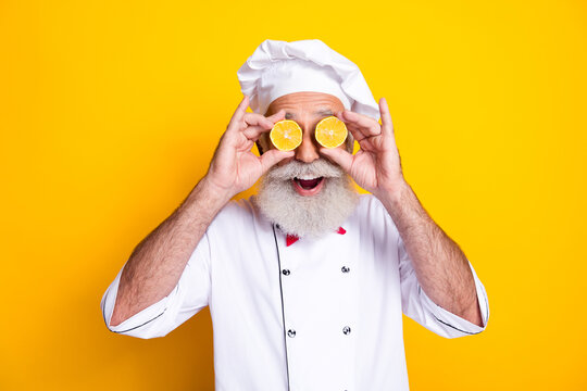 Cheerful senior chef holding lemon slices over eyes against vibrant yellow background
