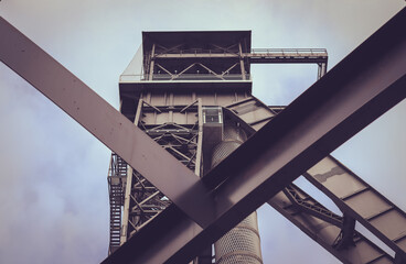 Naklejka premium An industrial structure, likely part of a factory or plant. The structure includes a tall metal tower with various platforms, ladders, and pipes. Large metal beams cross in front, forming an x shape.