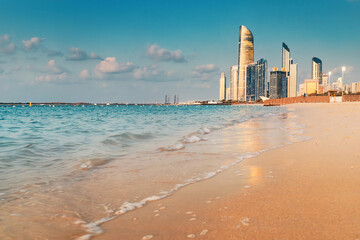 Beautiful beach with calm waves washing the sand at sunset, with modern skyscrapers of Abu Dhabi in United Arab Emirates