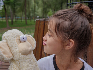 Cute girl with bun hairstyle plays ape and pouting lips to her funny toy stuffed monkey with bulging eyes and lips sitting on a bench in a city park outdoors.