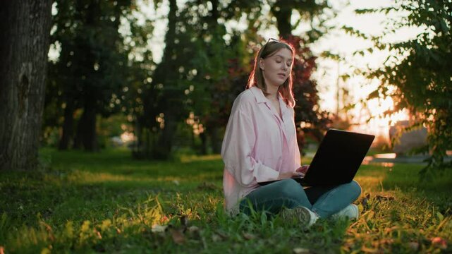 Young lady in pink top with glasses resting on her head, smiling warmly while typing on her laptop, she looks up thoughtfully, then refocuses back on her work as the sun creates a glowing atmosphere