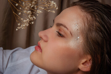Contemplative woman with glitter makeup and dried flowers in serene setting. Caucasian woman with...