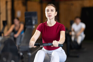 Focused girl works out with rowing machine during class at fitness club. Gym visitor performs exercise, creates load on muscles.