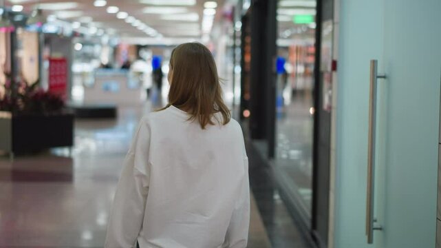 Back view of a shopper in a white top walking through a shopping mall, looking around amid colorful lights and a blurred background with unclear objects