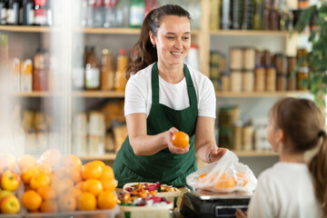 Fototapeta premium Buying ripe tangerines in store - female salesperson weighs tangerines on scales and hands them to a teenage girl