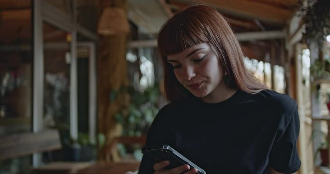 A young woman with auburn hair and bangs sits in a restaurant using a smart phone. She looks concerned as she reads and scrolls.