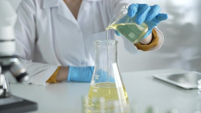 A scientist, wearing a lab coat and blue protective gloves, is pouring a yellow liquid from one beaker to another in laboratory, close up. Medicine and science concepts