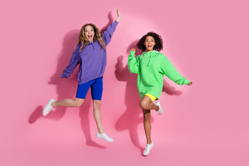 Two joyful young women jumping on a pink background wearing stylish hoodies and sportswear,...