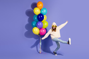 Young girl in casual pullover posing cheerfully with multicolored balloons against purple background