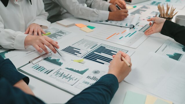 A collaborative business meeting scene depicting several hands engaged in analyzing financial reports and charts while discussing strategies and insights around a conference table. SACTR