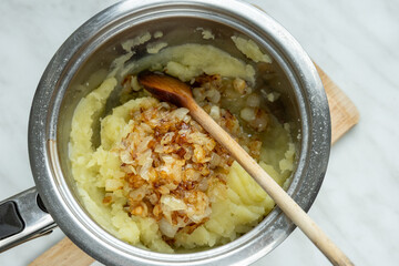 A stainless steel pot filled with mashed potatoes topped with caramelized onions sits on a wooden cutting board. 