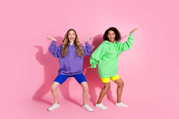 Energetic duo of young women posing together in colorful hoodies with expressive gestures on a vivid pink background