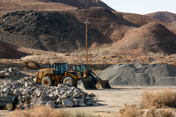 Two heavy duty front end loaders at a construction site with piles of gravel and boulders.