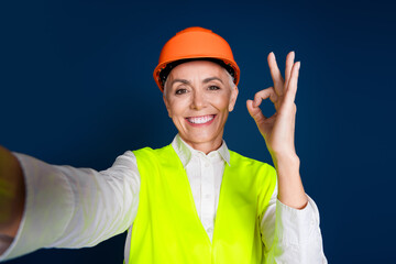 Confident senior woman engineer making OK gesture while wearing a high-visibility vest and hardhat...