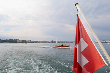 Swiss flag waving on a boat over Lake Geneva in Switzerland with the iconic Jet d'Eau fountain in the background under an overcast sky.