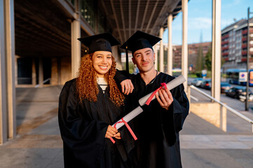 Two smiling students wearing graduation gowns and caps holding their diploma on their graduation day