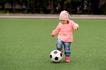 Obraz premium Little football player: little girl in pink jacket walking along green soccer field ready to kick ball. Young sportsman, future champion, healthy lifestyle, and active childhood concept. Copy space