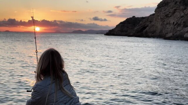 Blonde girl wearing down jacket fishing from rocks on the calm coast, with sun setting behind clouds in the background. Medium shot