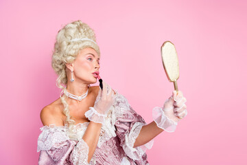 Elegant woman in historical royal costume applying lipstick while holding mirror on pink background