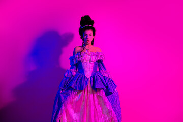 Elegant young woman in a baroque dress posing with neon ultraviolet lighting against a vibrant pink...