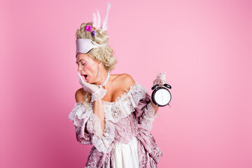 Fototapeta premium Elegant woman dressed in baroque costume yawning with vintage clock in hand against pink background