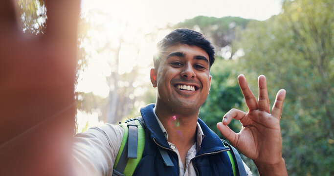 Selfie, vlogging or happy man hiking in nature with perfect sign, adventure or travel picture. Okay hand gesture, live streaming or Indian influencer in forest for social media photograph or journey