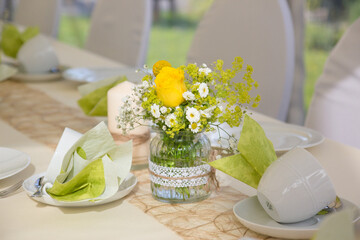 Beautiful wedding table setup on a sunny day with light green napkins, yellow roses, and daisies in lace-decorated vases, creating a romantic and fresh ambiance.