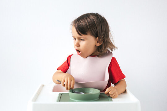 Little girl sitting in a high chair, wearing a pink bib and red shirt, coughing while eating. She holds a spoon in one hand and looks uncomfortable, possibly choking on food during her meal