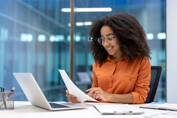 Successful businesswoman at workplace inside office working with documents, reports and contracts. Woman smiling happily, using laptop at work.