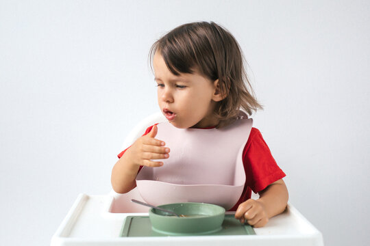 Little girl sitting in high chair, wearing pink bib and red shirt, coughing while eating. holds a spoon in one hand, with food on her lips, and touches her chest, showing discomfort, choking on meal