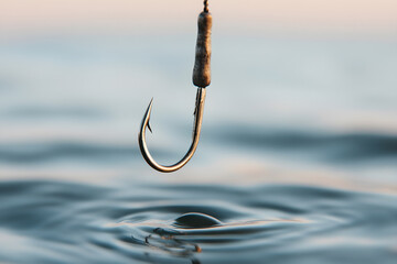 A fishing hook descends toward the surface of a body of water, poised to enter. The hook, still shiny, reflects the sky and contrasts with the water's surface.