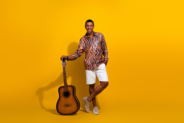 Young man with guitar poses in front of a vivid yellow background in summer casual attire