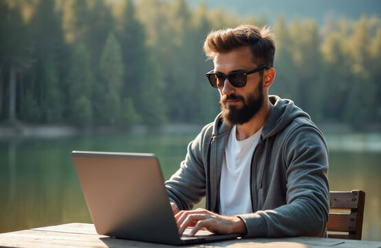 Bearded man works on laptop at wooden table against lake, forest backdrop. Digital nomad uses computer, enjoys remote work, outdoor office. Freelancer types on keyboard, stays connected.