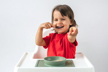 Smiling little girl in a red shirt sitting in a high chair, eating soup with a spoon and playfully...