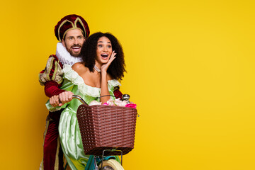 Couple in elegant masquerade costumes riding a vintage bicycle on a bright yellow background