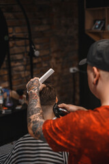 A barber man with a tattoo on his arm makes a haircut to a brunette guy in a barbershop, cuts his hair with a clipper close-up