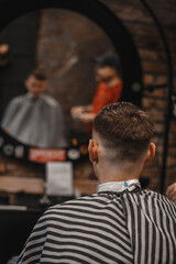 A barber man with a tattoo on his arm makes a haircut to a brunette guy in a barbershop, cuts his hair with a clipper close-up