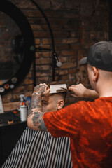 A barber man with a tattoo on his arm makes a haircut to a brunette guy in a barbershop, cuts his hair with a clipper close-up