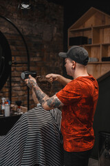 A barber man with a tattoo on his arm makes a haircut to a brunette guy in a barbershop, cuts his hair with a clipper close-up